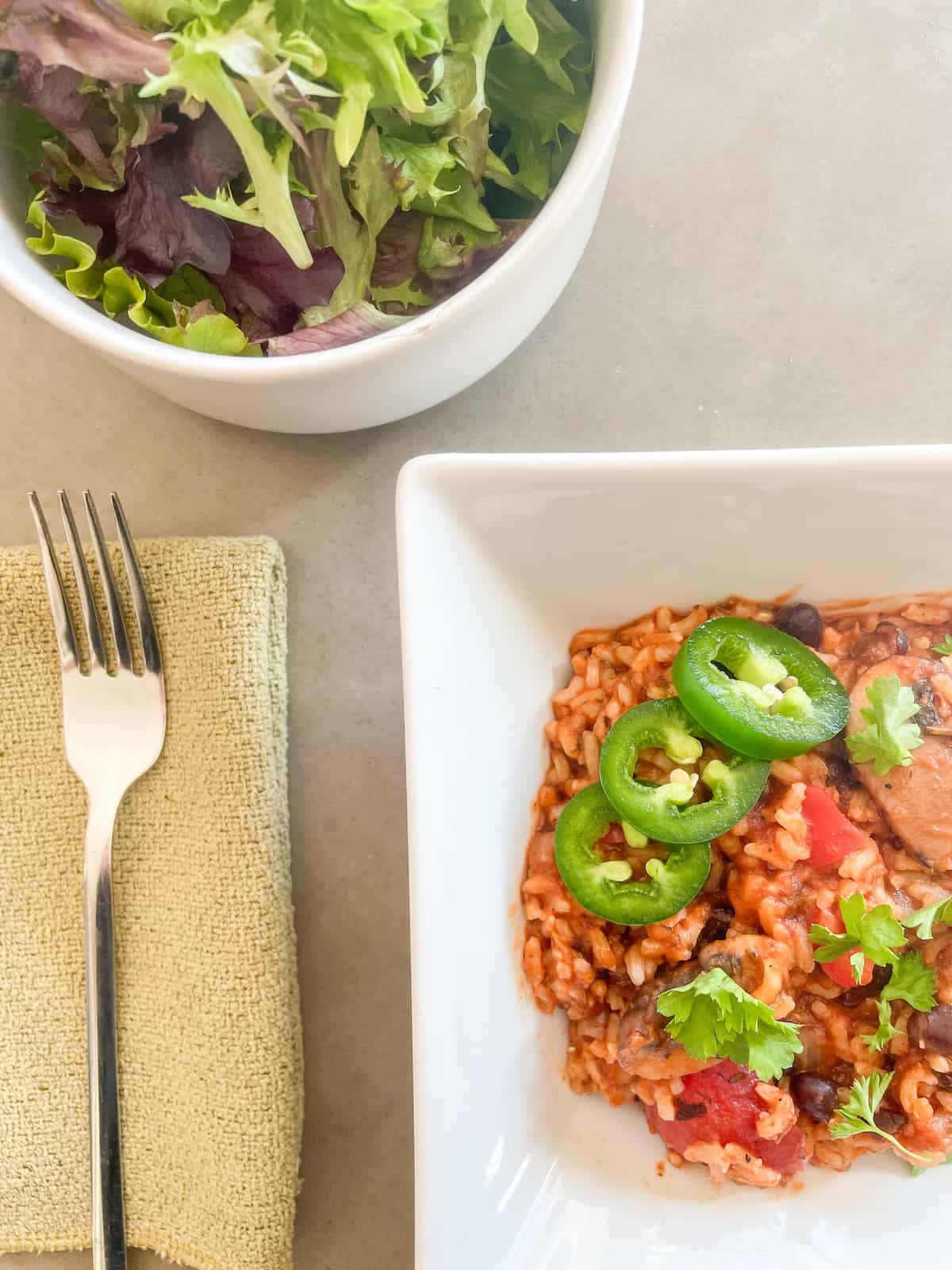 Bowl of Black Bean Jambalaya topped with Jalapeno peppers and parsley. Side salad and a fork.