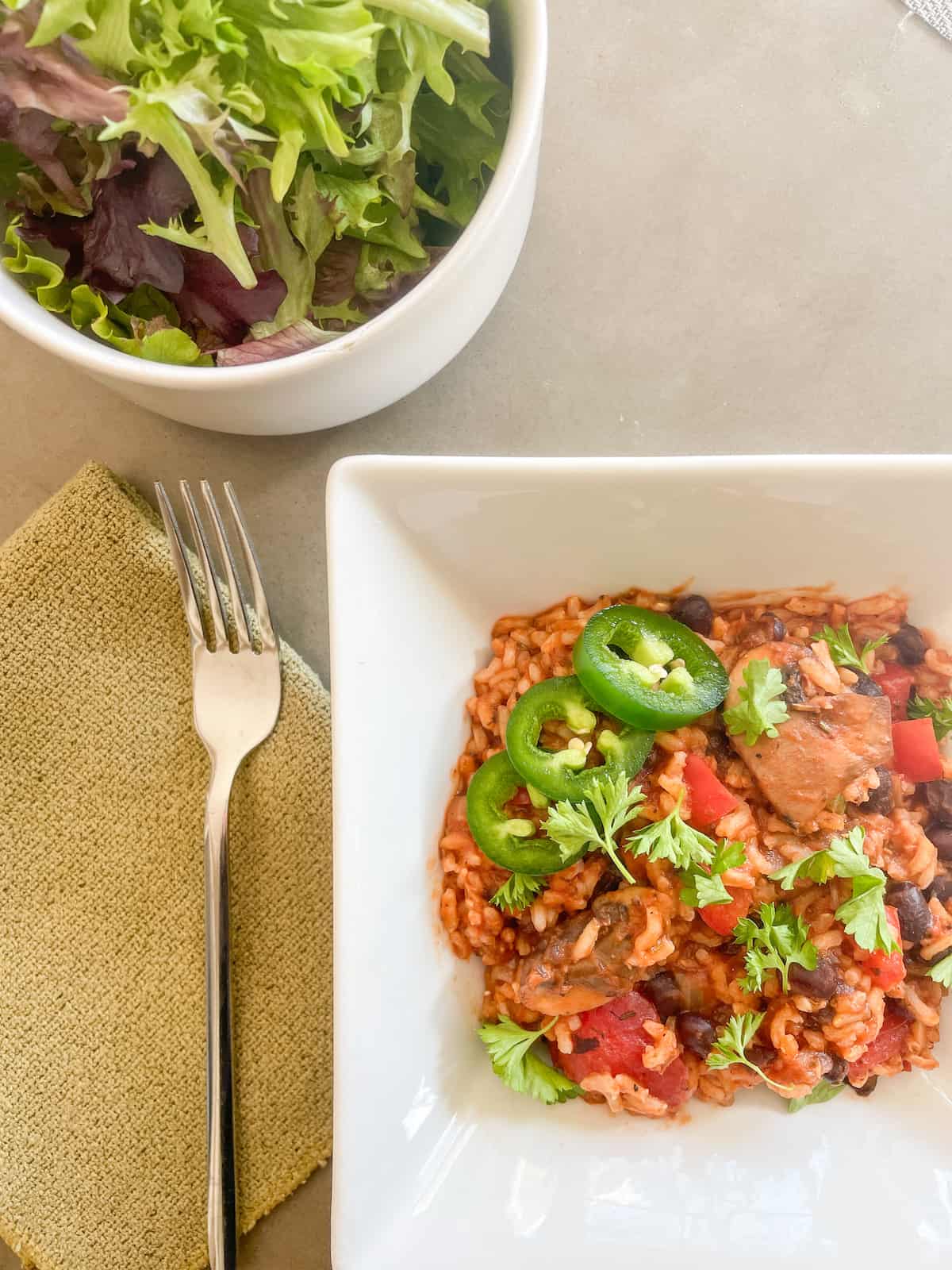 Bowl of Black Bean Jambalaya topped with Jalapeno peppers and parsley. Side salad and a fork.